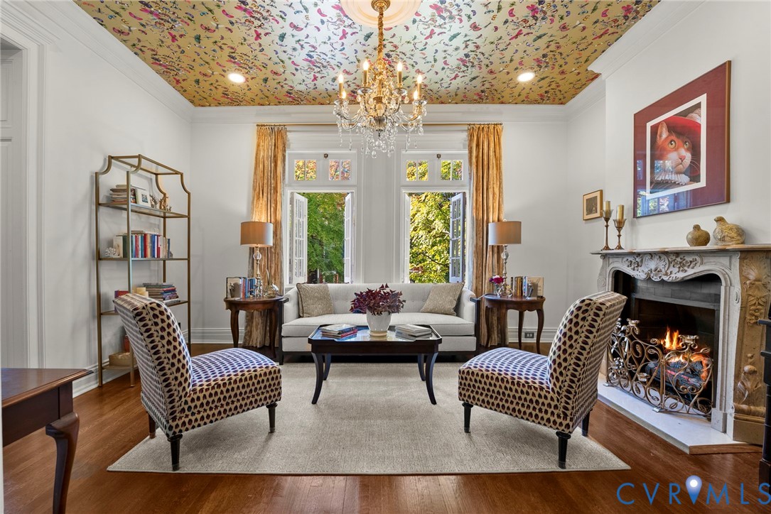Living space featuring patterned ceiling wallpaper, ornate crystal chandelier, hardwood flooring, and a decorative carved fireplace surround with a metal hearth screen