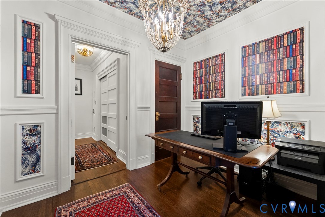 Intricate crown molding and wall paneling, elaborate patterned ceiling wallpaper, polished wood-finish flooring, solid wood five-panel door, and a multi-tiered crystal chandelier