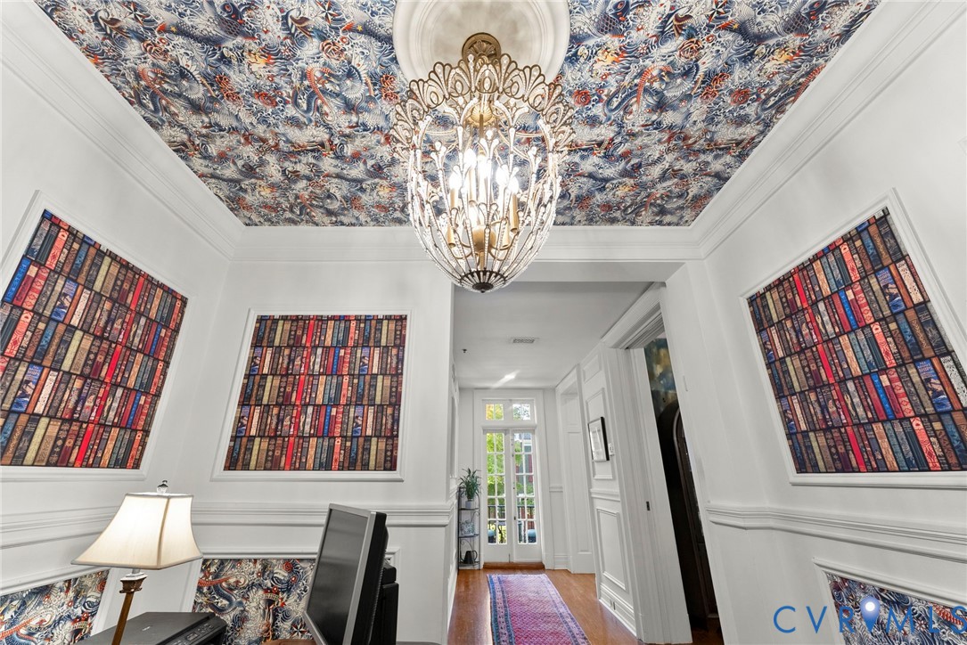 Intricate patterned ceiling wallpaper with matching wall panel inserts, ornate metal chandelier, central ceiling medallion, and white wainscoting