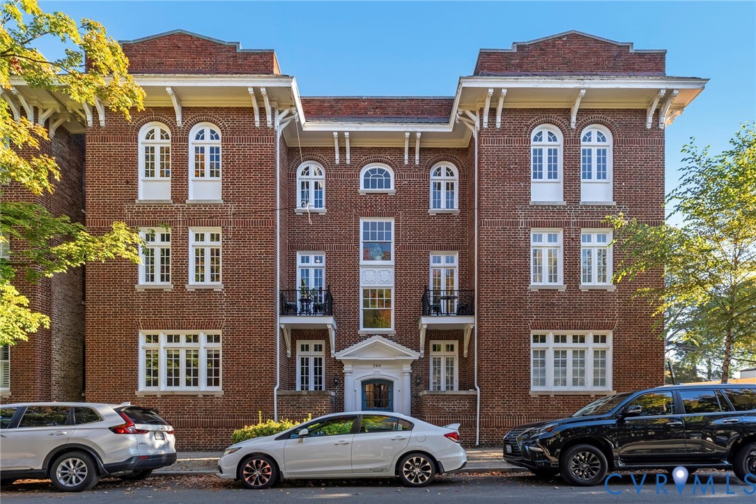 Brick facade exterior featuring arched window detailing, overhanging eaves with decorative brackets, two wrought-iron balconies, and a formal entryway pediment