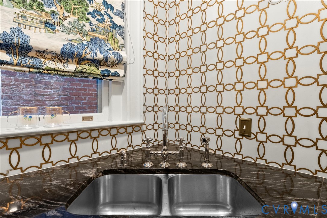 Double-basin stainless steel sink with a polished metal bridge-style faucet and side sprayer, set into a dark natural-stone countertop with integrated backsplash, complemented by geometric-patterned wall covering