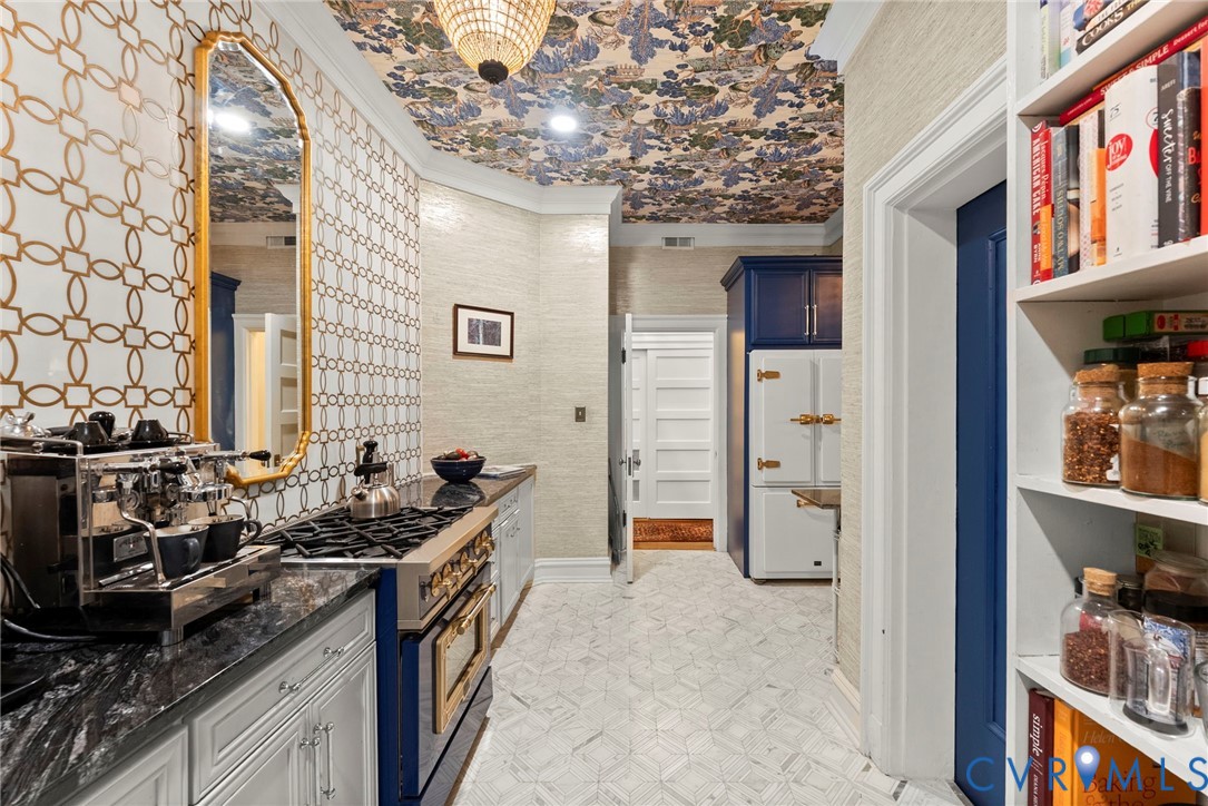 Patterned wallpaper ceiling, custom blue cabinetry with metallic hardware, geometric tile backsplash, stone countertop, and built-in shelving