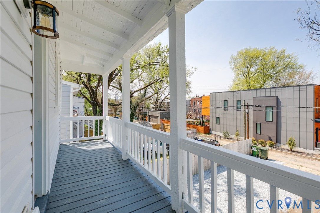 Covered porch featuring wood-finish decking, white railings, structural support columns, and an exterior wall-mounted light fixture