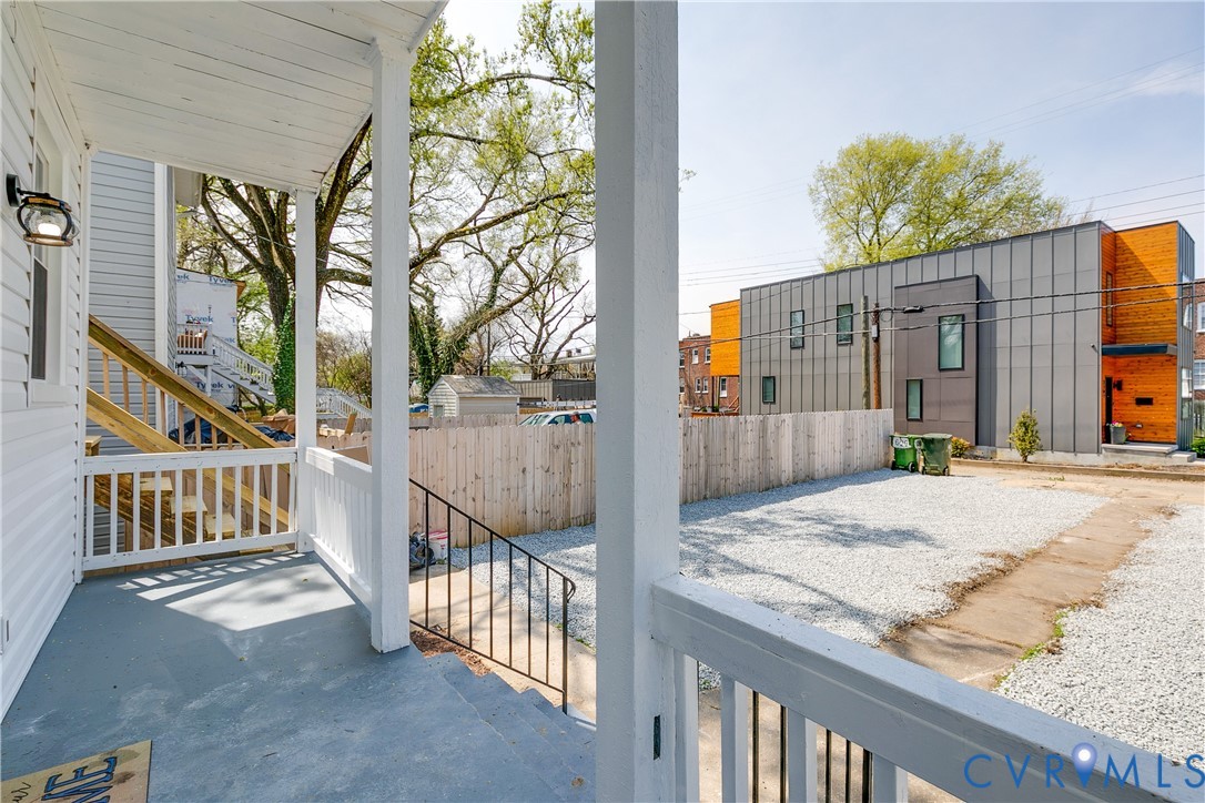 Covered front porch with white railing, exterior wall-mounted light fixture, gravel parking area, and wood privacy fencing