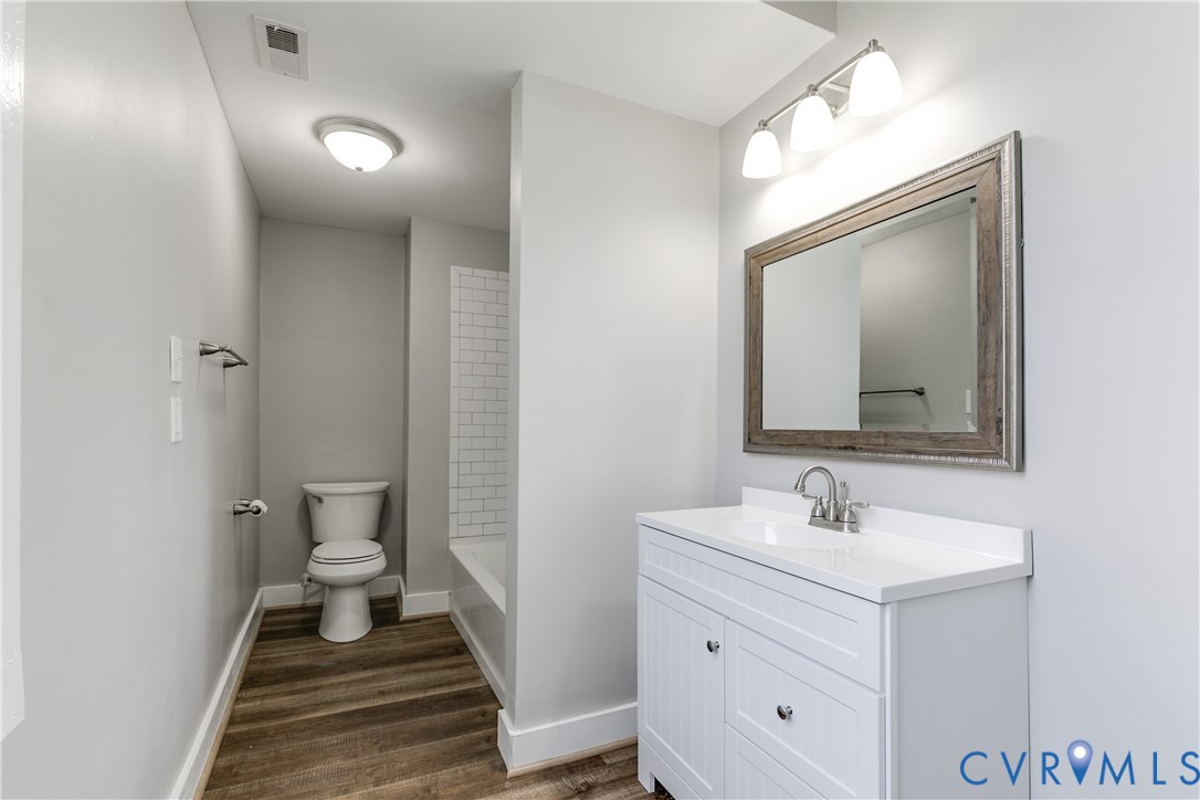 White vanity cabinet featuring a solid surface countertop, framed mirror, three-light fixture, wood-finish flooring, and tub surround with white subway tile
