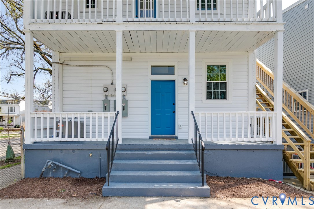 Exterior featuring dual-level covered porches with white railing, blue paneled entrance door, single-hung window with transom, and wooden staircase