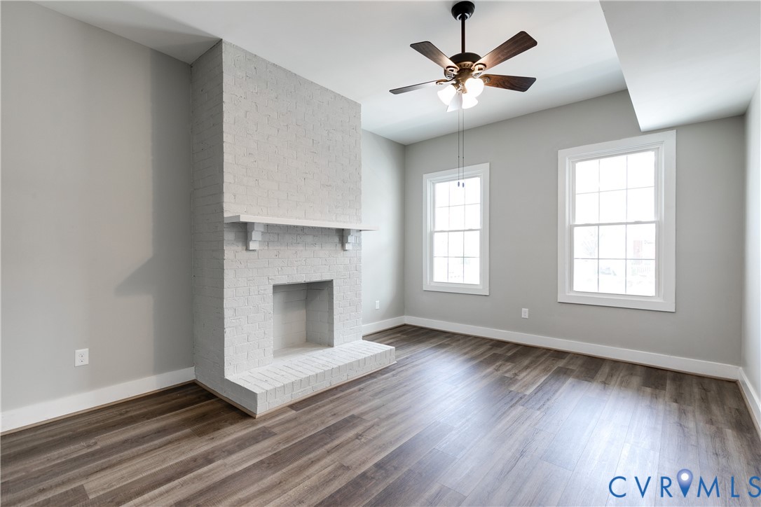 Painted brick fireplace with a mantel, wood-finish flooring, two multi-pane windows, and a ceiling fan with light fixture