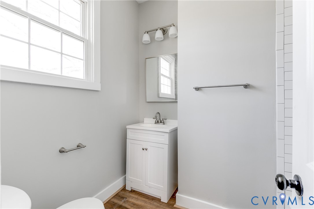 White vanity with a neutral solid surface countertop, rectangular under-mount sink, three-light polished chrome vanity fixture, framed wall mirror, and wood-finish flooring