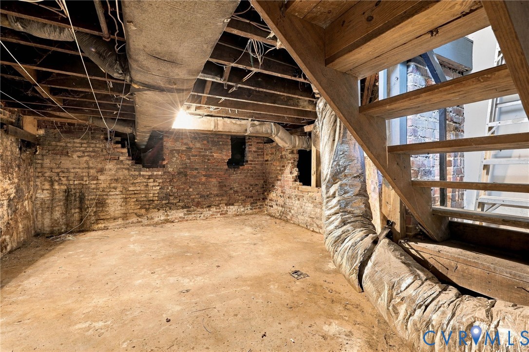 Unfinished lower level featuring exposed wooden ceiling joists, brick foundation walls, and an open wooden staircase