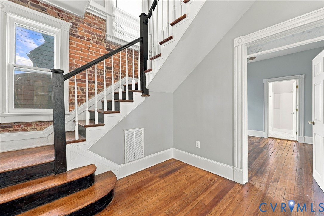 Exposed brick accent wall, wood-finish flooring, and a staircase featuring wood treads with a dark-finished handrail and white balusters Exposed brick accent wall, wood-finish flooring, and a staircase featuring wood treads with a dark-finished handrail and white balusters