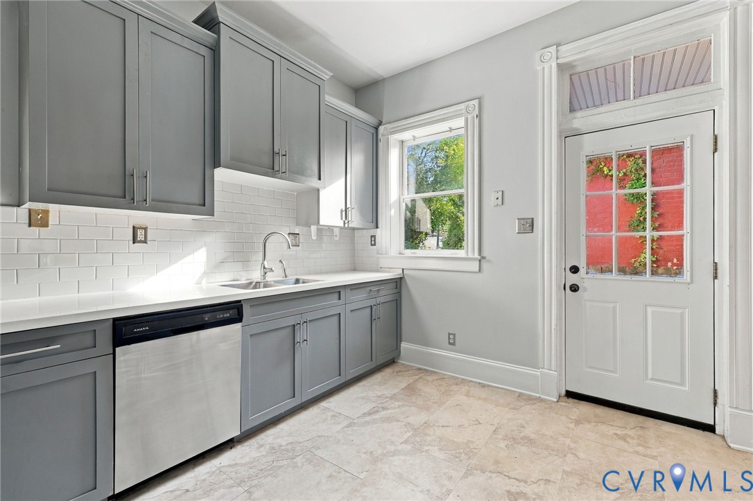 Kitchen featuring gray shaker-style cabinetry, white subway tile backsplash, solid surface countertops, stainless steel dishwasher, and stone-finish tile flooring