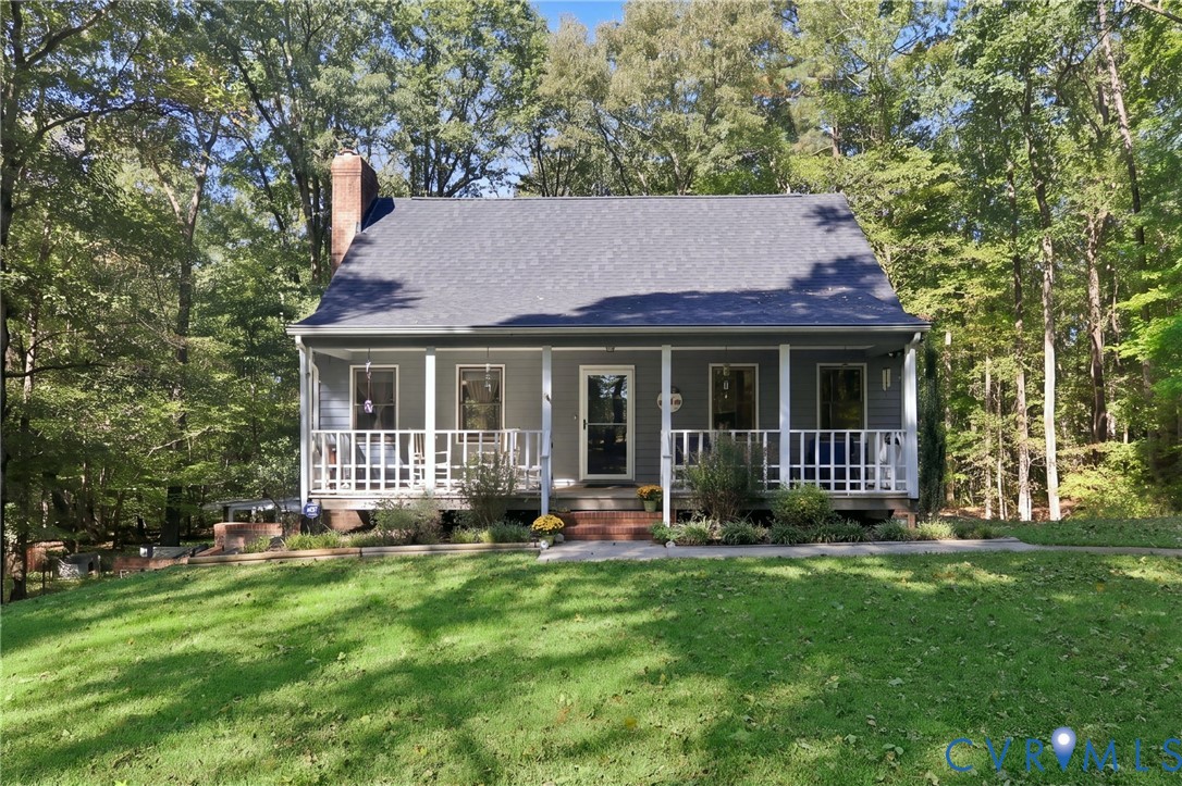 Cape Cod style exterior featuring a covered front porch with railings, a brick chimney, and a backdrop of mature trees Cape Cod style exterior featuring a covered front porch with railings, a brick chimney, and a backdrop of mature trees