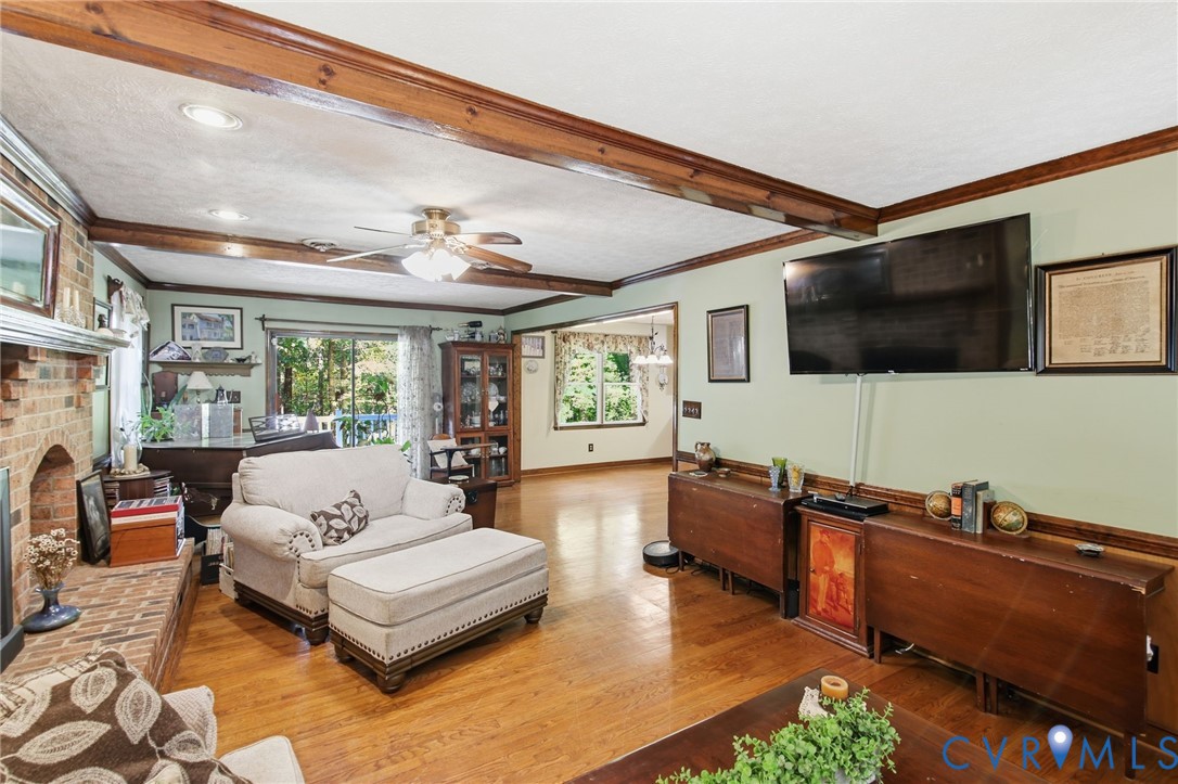 Living area featuring exposed stained wood ceiling beams, brick fireplace surround, wood-finish flooring, and crown molding Living area featuring exposed stained wood ceiling beams, brick fireplace surround, wood-finish flooring, and crown molding
