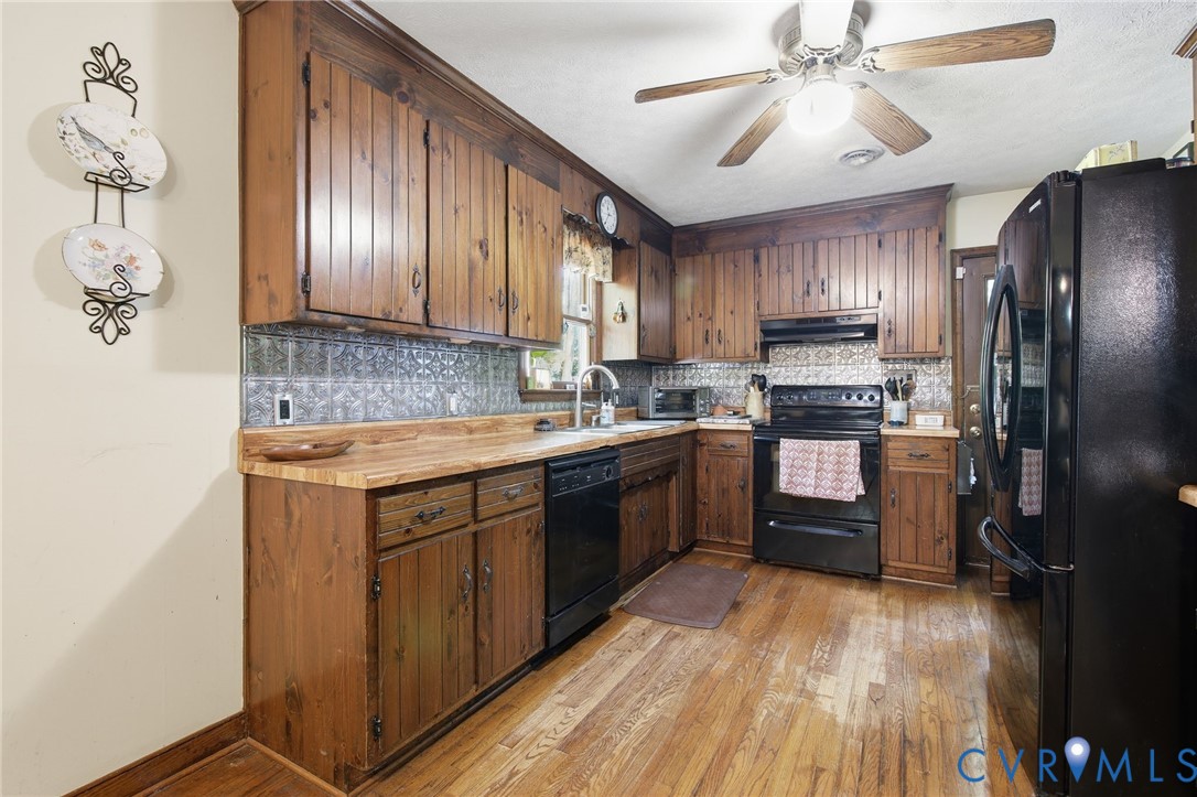 Kitchen featuring custom wood cabinetry, a decorative metallic tile backsplash, butcher block countertops, black appliances, and wood flooring