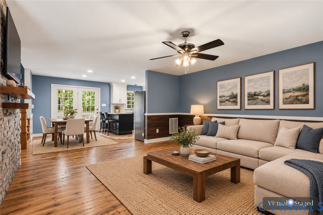 Wood-finish flooring, floor-to-ceiling stone-veneer fireplace, wood-panel wainscoting, glass-paneled double doors, and ceiling fan with integrated lighting Wood-finish flooring, floor-to-ceiling stone-veneer fireplace, wood-panel wainscoting, glass-paneled double doors, and ceiling fan with integrated lighting