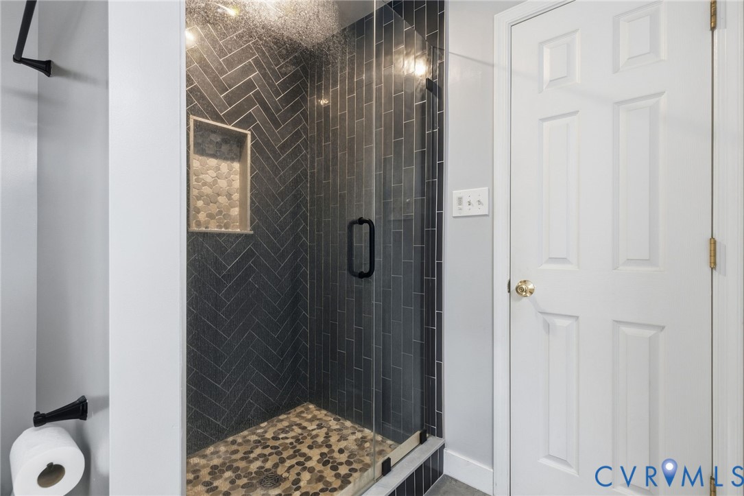 Walk-in shower featuring dark herringbone tile walls, a built-in recessed niche, pebble-stone flooring, and a frameless glass door with matte black hardware