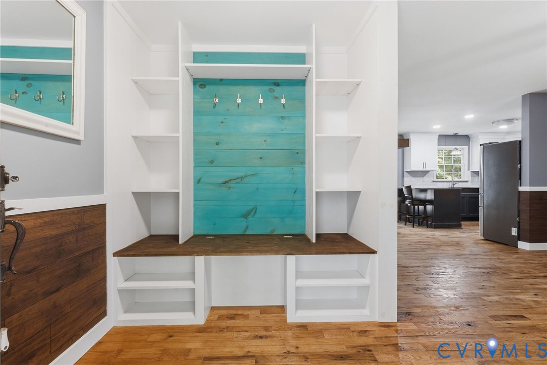 Built-in mudroom storage featuring white shelving, a stained wood bench, blue horizontal plank backing, and metal coat hooks, complemented by wood-finish flooring and wainscoting