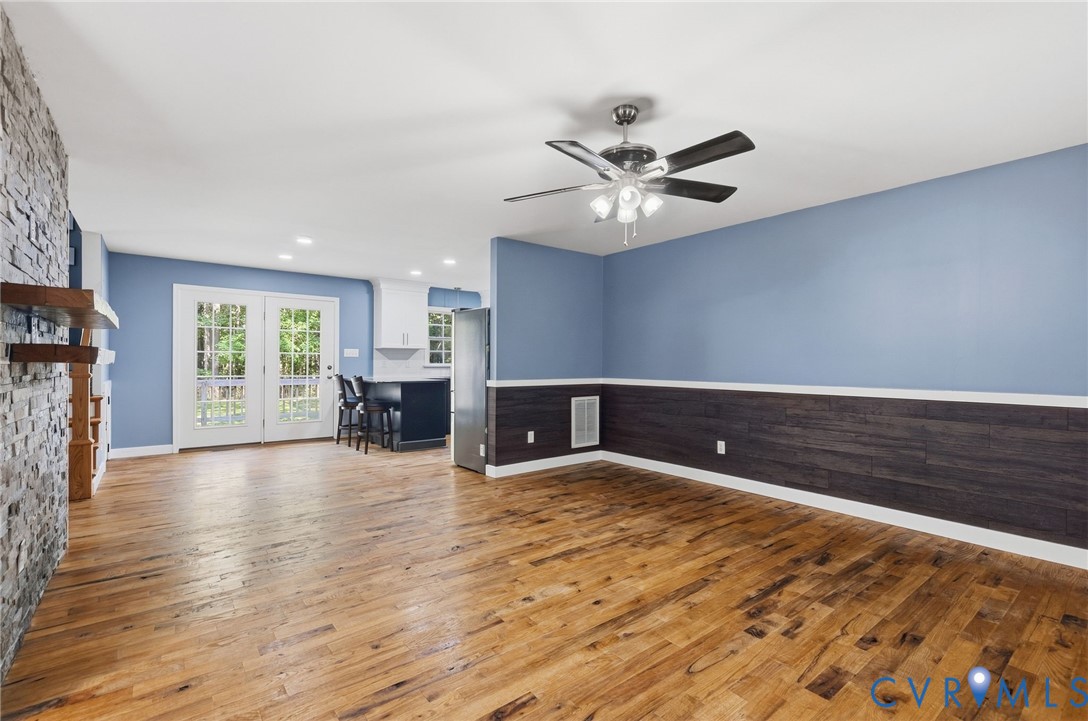 Wood-finish flooring, stone-clad fireplace, dark wood-tone wainscoting, ceiling fan with integrated lighting, and multi-pane French doors