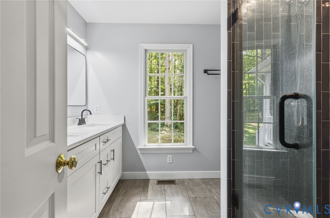 Bathroom featuring a double-sink vanity with white cabinetry and solid-surface countertop, walk-in shower with dark tile surround and glass door, and wood-finish tile flooring