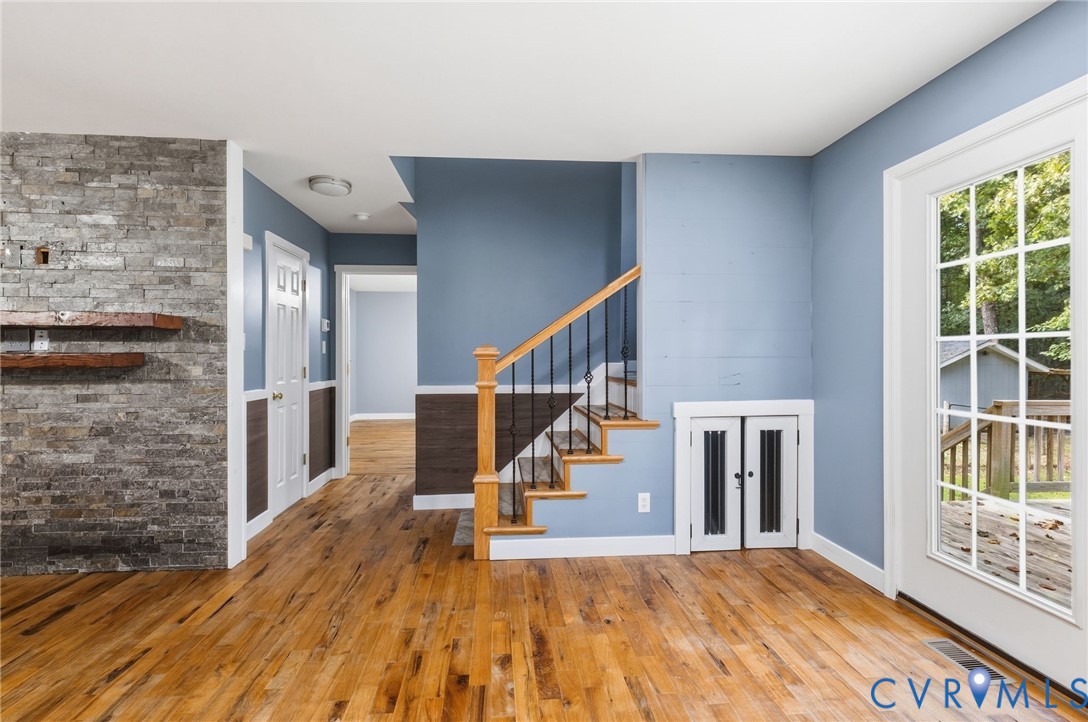 Interior featuring wood-finish flooring, a full-height stone-veneer fireplace with wood-beam mantels, a staircase with decorative metal spindles, and a multi-pane glass door