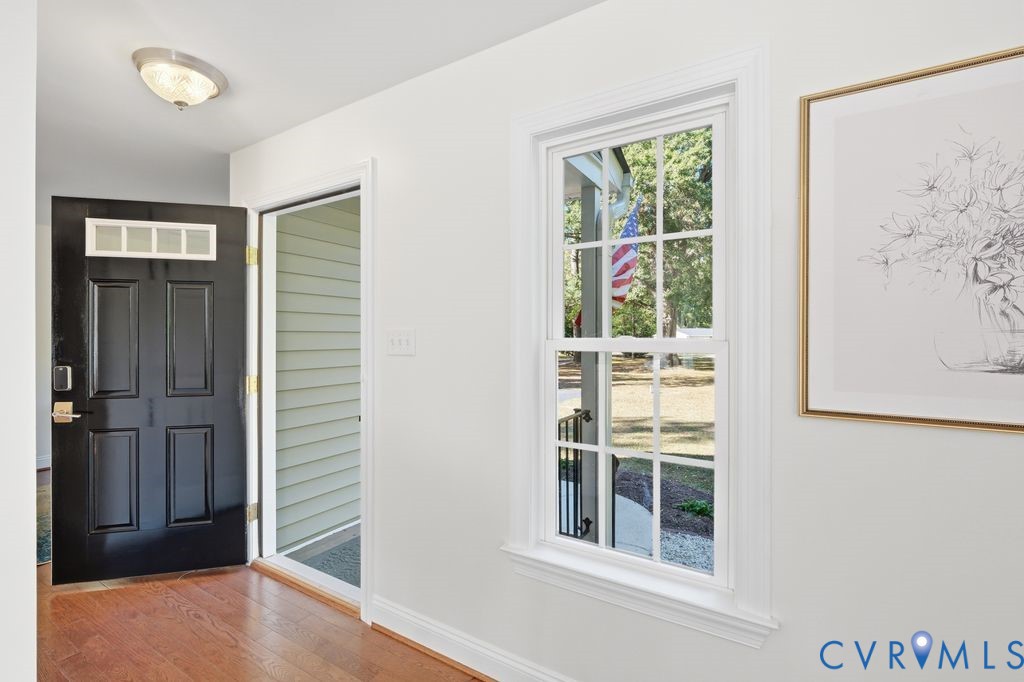 Interior hallway featuring wood-finish flooring, a black paneled door with multi-pane transom lites, extensive white trim, a multi-pane double-hung window, and a flush-mount ceiling light fixture