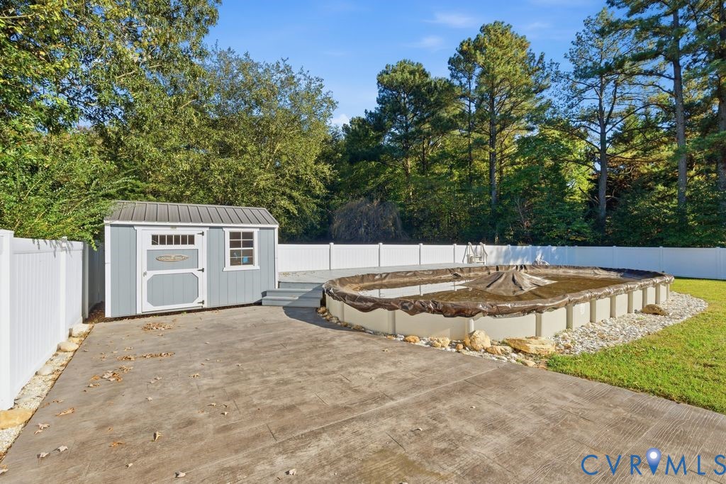 Backyard featuring an above-ground pool, concrete patio, detached storage structure with a metal roof, and white vinyl perimeter fencing