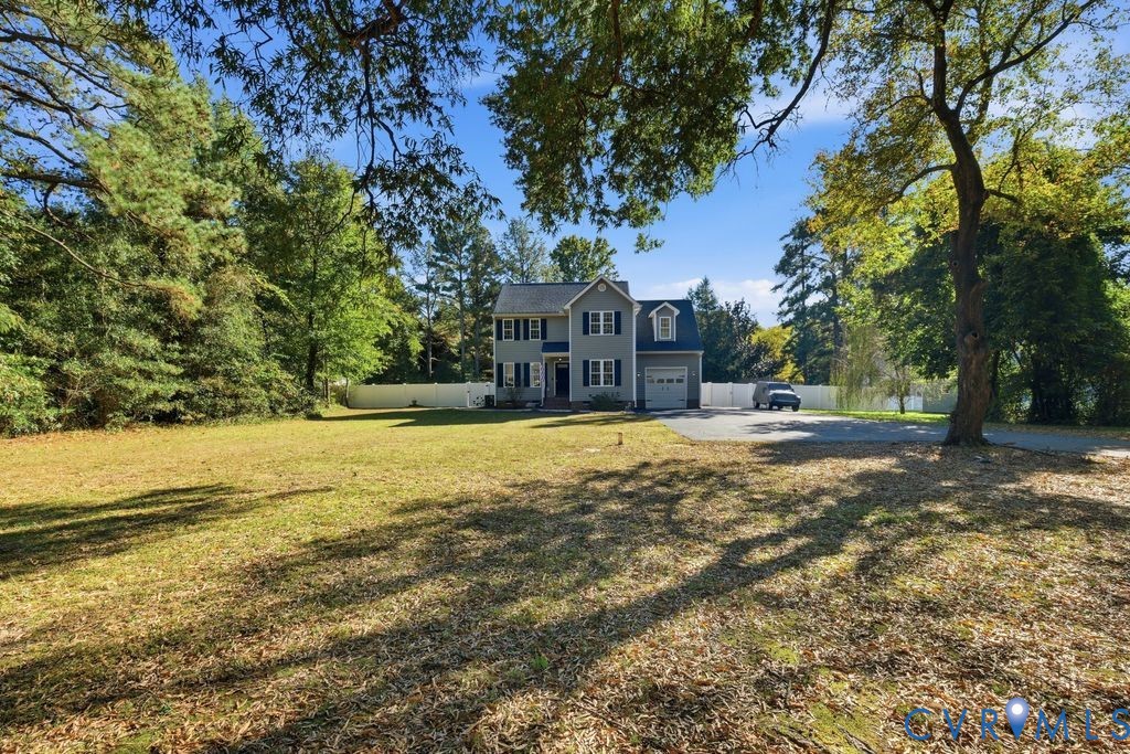Two-story residence featuring light-toned siding, dark window shutters, an attached garage, a paved driveway, and a white privacy fence