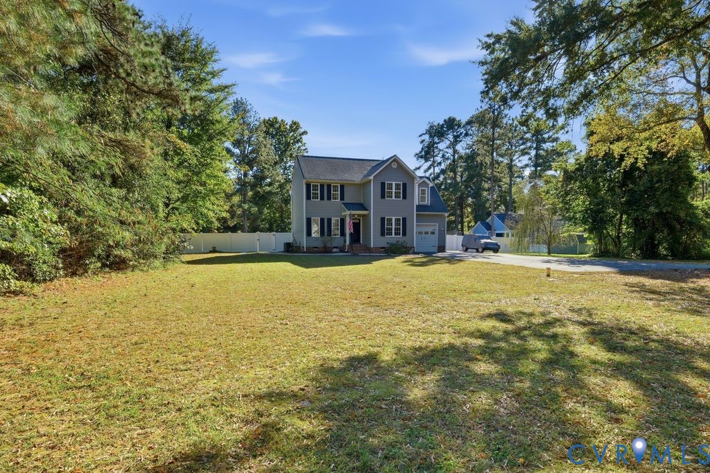 Two-story residence featuring a gray siding exterior, attached one-car garage, white vinyl fencing, and an expansive lawn with mature trees