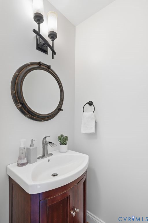 Wood-finish pedestal vanity with a white basin, metallic faucet, circular porthole-style wall mirror, and dual-light sconce