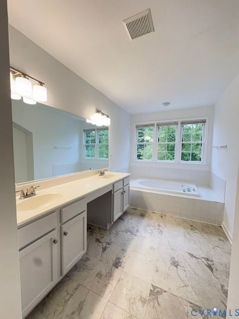 Dual-sink vanity with neutral cabinetry, expansive wall-to-wall mirror, tiled soaking tub, and patterned stone-finish flooring Dual-sink vanity with neutral cabinetry, expansive wall-to-wall mirror, tiled soaking tub, and patterned stone-finish flooring