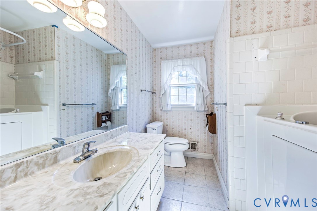 Full-width vanity with stone-finish countertop, integrated sink, expansive wall-mounted mirror, ceramic tile flooring, and tub-shower combination with ceramic wall surround