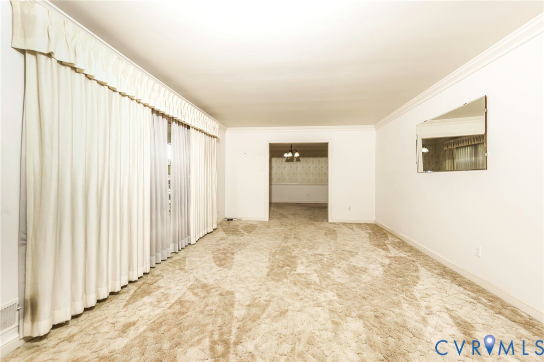 Spacious room featuring floor-to-ceiling drapery, neutral carpeting, crown molding, and a mounted wall mirror Spacious room featuring floor-to-ceiling drapery, neutral carpeting, crown molding, and a mounted wall mirror