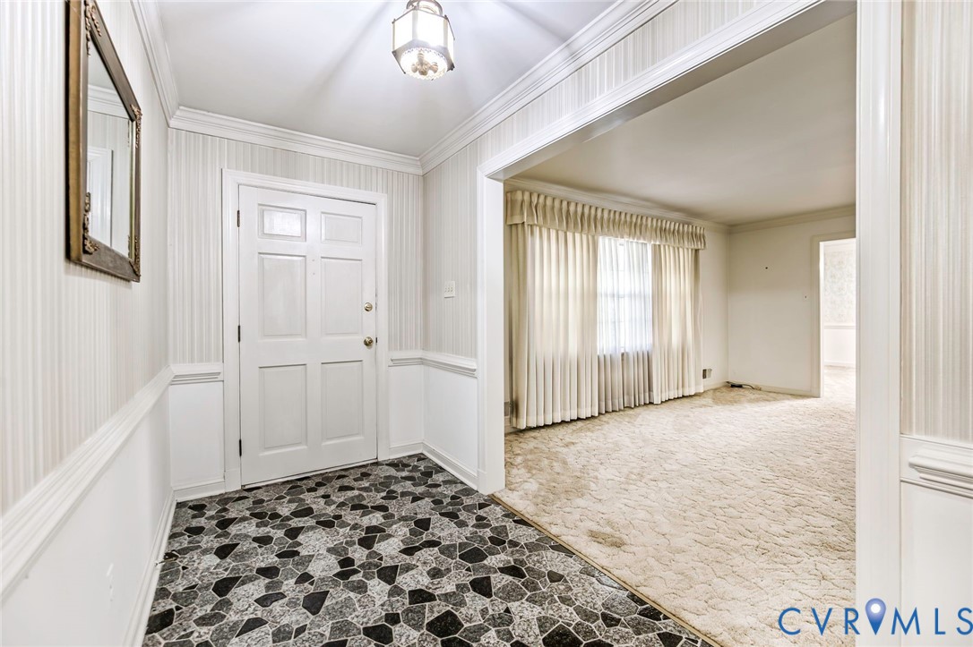 Foyer featuring stone-patterned tile flooring, chair rail molding, wallpapered walls, crown molding, and a built-in overhead light fixture Foyer featuring stone-patterned tile flooring, chair rail molding, wallpapered walls, crown molding, and a built-in overhead light fixture
