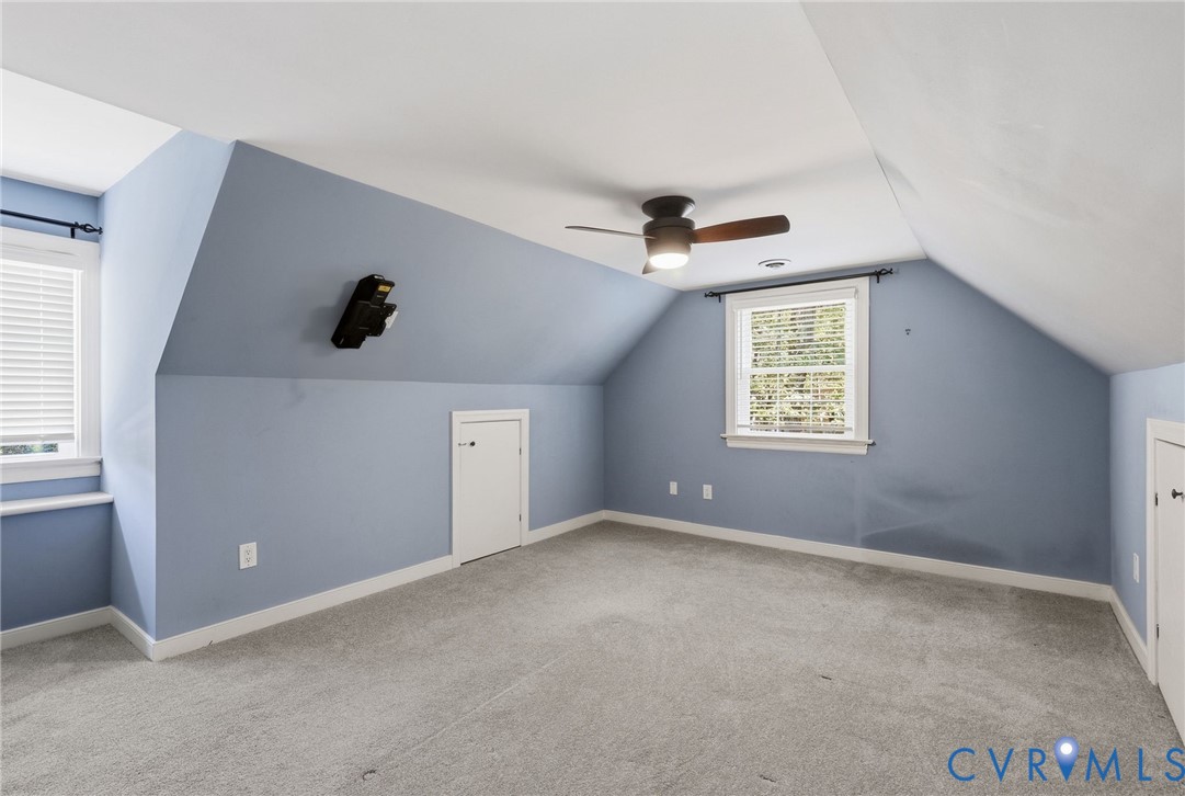 Room featuring sloped ceilings, carpeted flooring, two built-in knee-wall storage access doors, two windows, and an integrated ceiling fan with light fixture