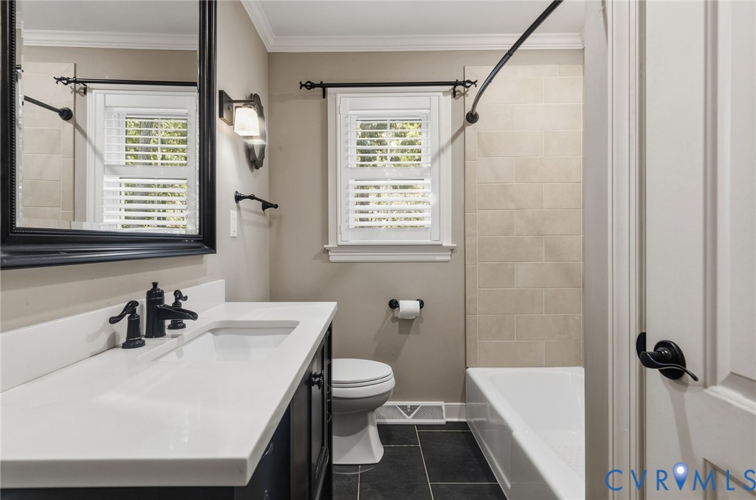 Bathroom featuring a white rectangular vanity with a solid surface countertop, matte black faucet, framed vanity mirror, dark floor tiling, and a tub enclosure with neutral wall tile