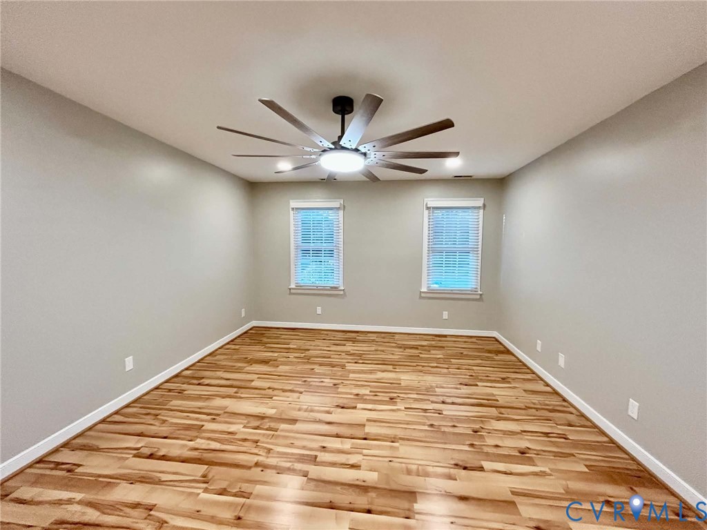 Neutral-toned room featuring wood-finish flooring, dual windows with integrated blinds, and a multi-blade ceiling fan with centered lighting