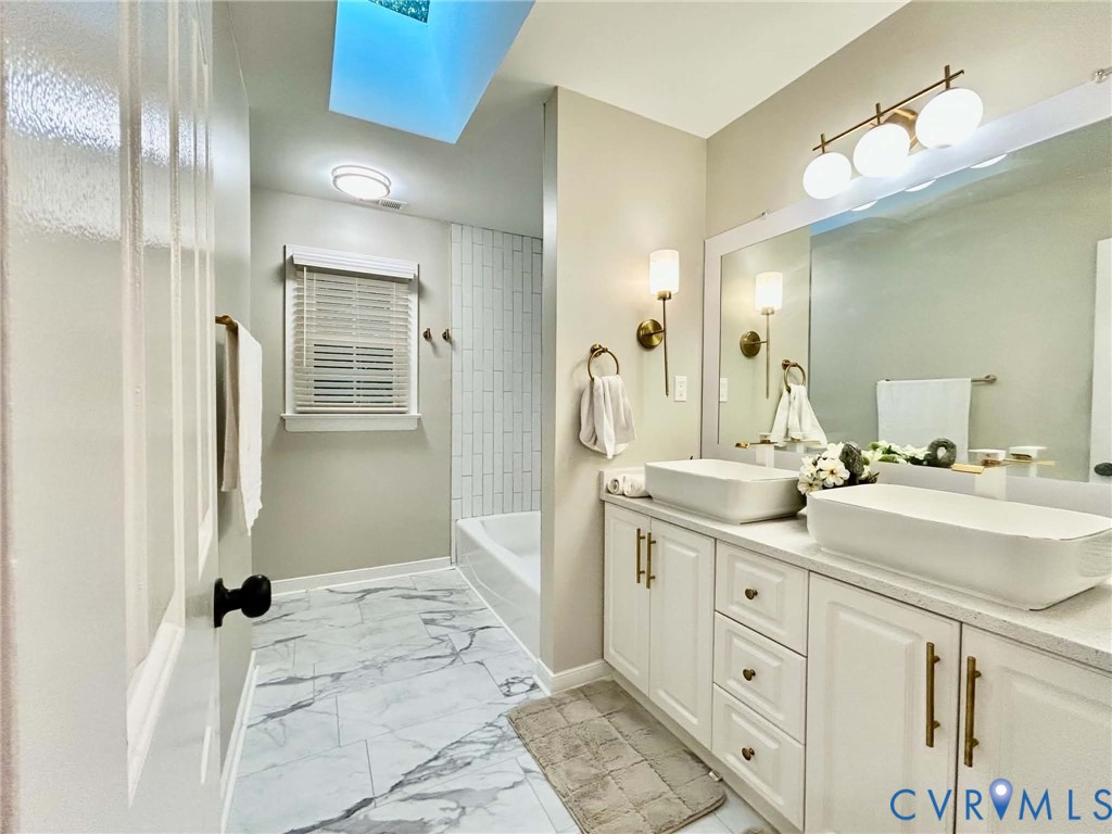 Double vanity with rectangular vessel sinks and quartz countertop, skylight, white cabinetry with brass-finish hardware, and tiled tub surround
