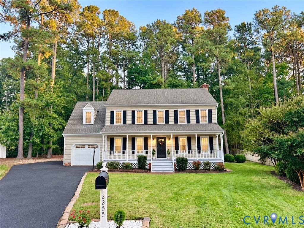 Colonial-style exterior featuring light-toned siding, a covered front porch with a white railing, black shutter accents, an attached garage, and a mature tree-lined perimeter