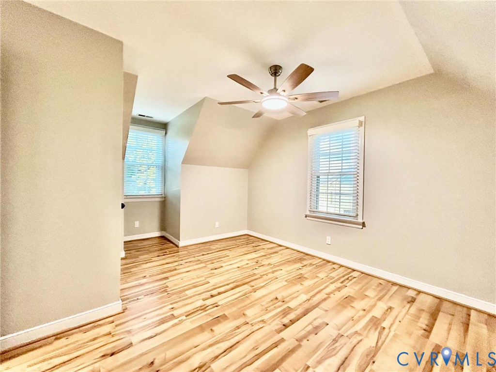 Vaulted ceiling featuring a central multi-blade ceiling fan with integrated lighting, wood-finish flooring, and dual windows with installed blinds