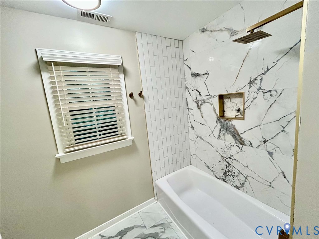 Bathtub surround featuring large-format stone-patterned tile, a recessed niche, vertical subway tile accents, and a gold-tone rainfall showerhead