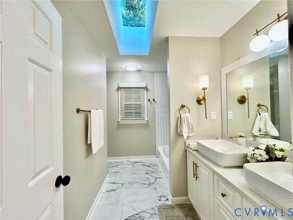 Bathroom featuring a skylight, dual vessel sinks on a quartz vanity, patterned tile flooring, and metallic wall sconces