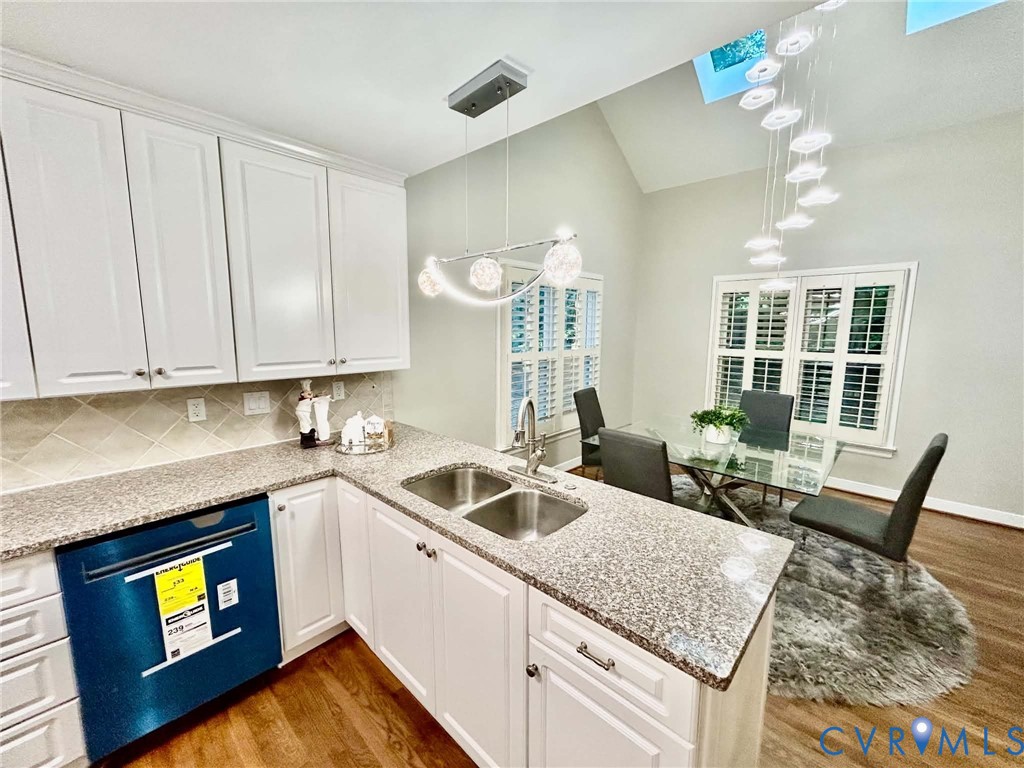 Kitchen featuring white shaker-style cabinetry, stone-finish countertops, stainless steel dual-basin undermount sink, vaulted ceiling with integrated skylights, and polished metal pendant lighting