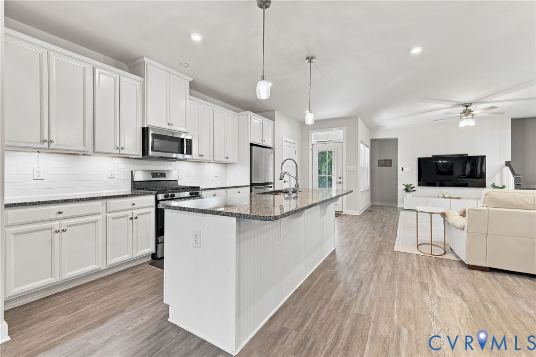 Kitchen featuring white shaker-style cabinetry, stone-surface countertops, a center island with a stainless steel faucet, subway tile backsplash, and wood-finish flooring Kitchen featuring white shaker-style cabinetry, stone-surface countertops, a center island with a stainless steel faucet, subway tile backsplash, and wood-finish flooring