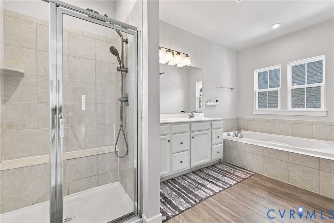 Bathroom featuring a tiled glass-enclosed shower, soaking tub with tile surround, white cabinetry, wood-finish flooring, and multi-light vanity fixture