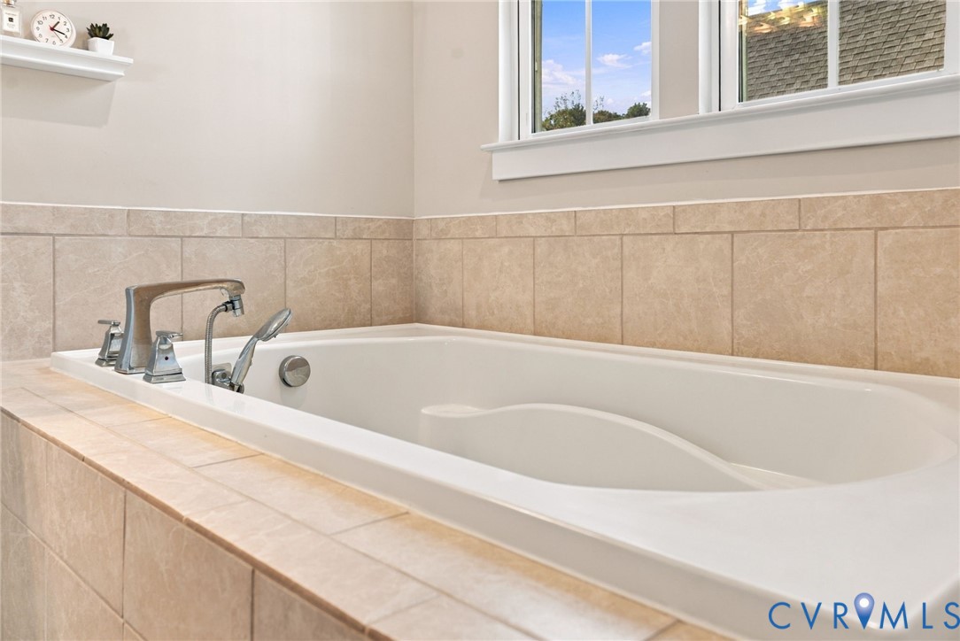 Soaking tub featuring a tiled surround, deck-mounted chrome faucet with handheld shower attachment, and integrated wall-mounted shelving