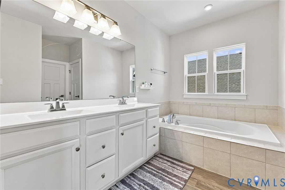 Double vanity with white cabinetry, solid surface countertop, inset soaking tub with tile surround, and large wall-mounted mirror