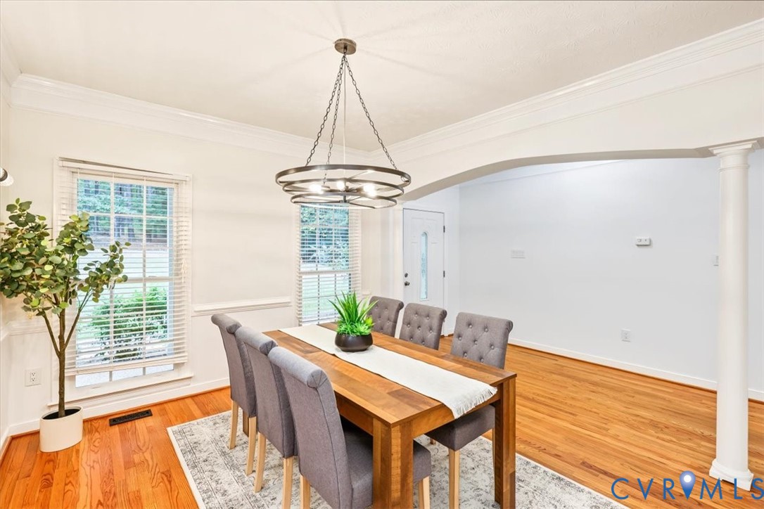 Dining area featuring wood-finish flooring, crown molding, a circular tiered chandelier, a structural arched opening supported by a column, and white wainscoting