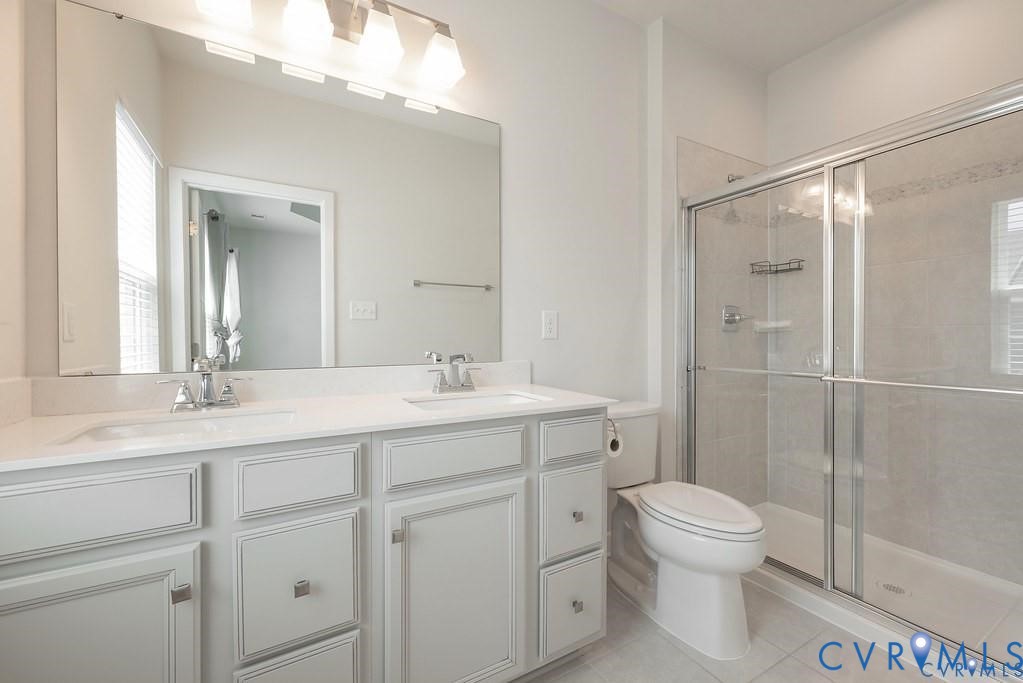 Bathroom featuring a dual-sink vanity with light-toned cabinetry, solid-surface countertops, a full-width mirror, and a glass-enclosed shower with tile surround