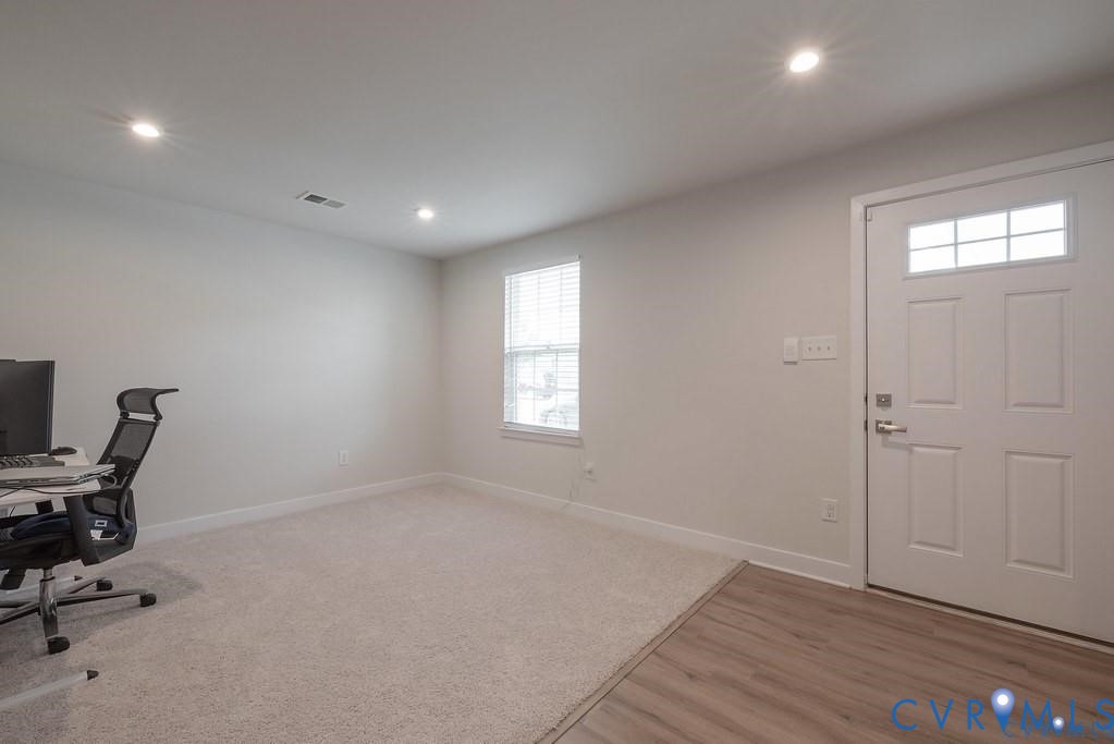 Neutral interior room featuring recessed lighting, carpeted flooring, wood-finish entryway flooring, and a six-panel exterior door with a windowed transom