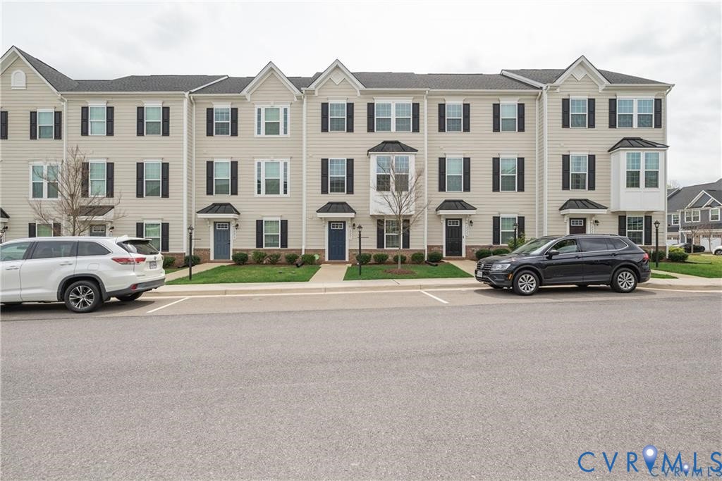 Multi-story townhome exterior featuring neutral vinyl siding, dark shutters, bay windows, gabled rooflines, and individual covered entryways